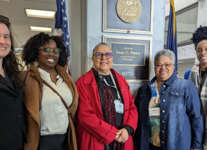 Five women smiling and standing together in front of Senator Gary C. Peters’ office sign at the U.S. Capitol. The sign reads “Senator Gary C. Peters, Michigan.” They are dressed in professional attire and appear to be visiting as part of a delegation.
